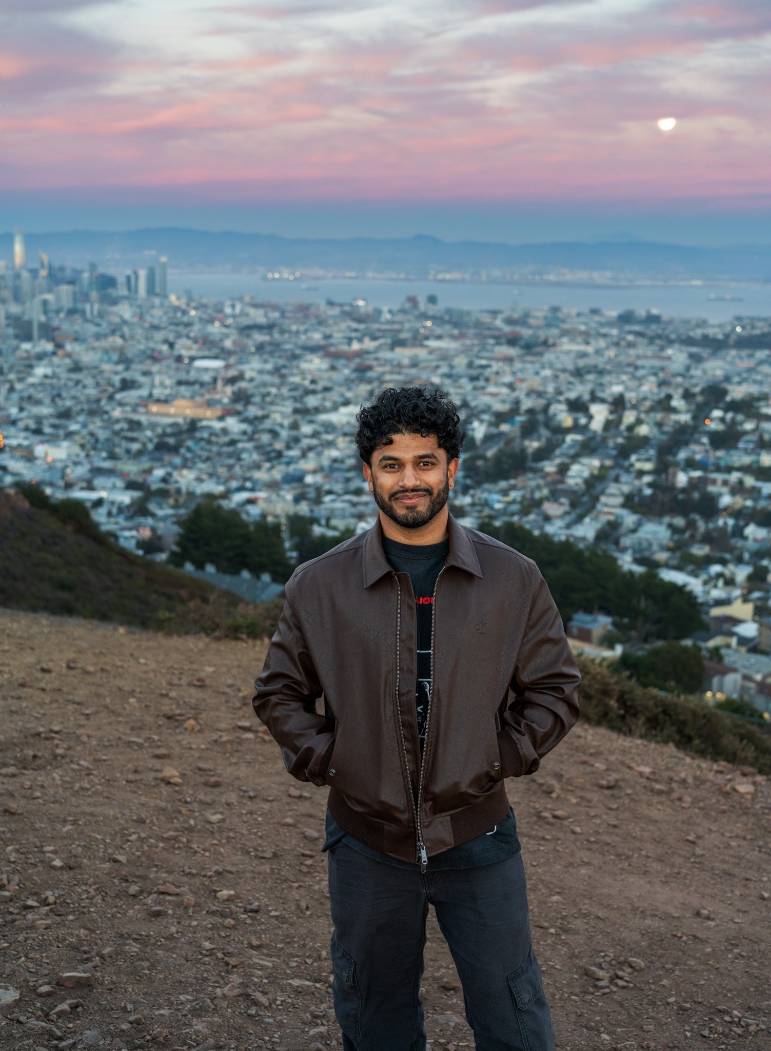Harshith smiling in a brown jacket, with the city behind him at sunset
