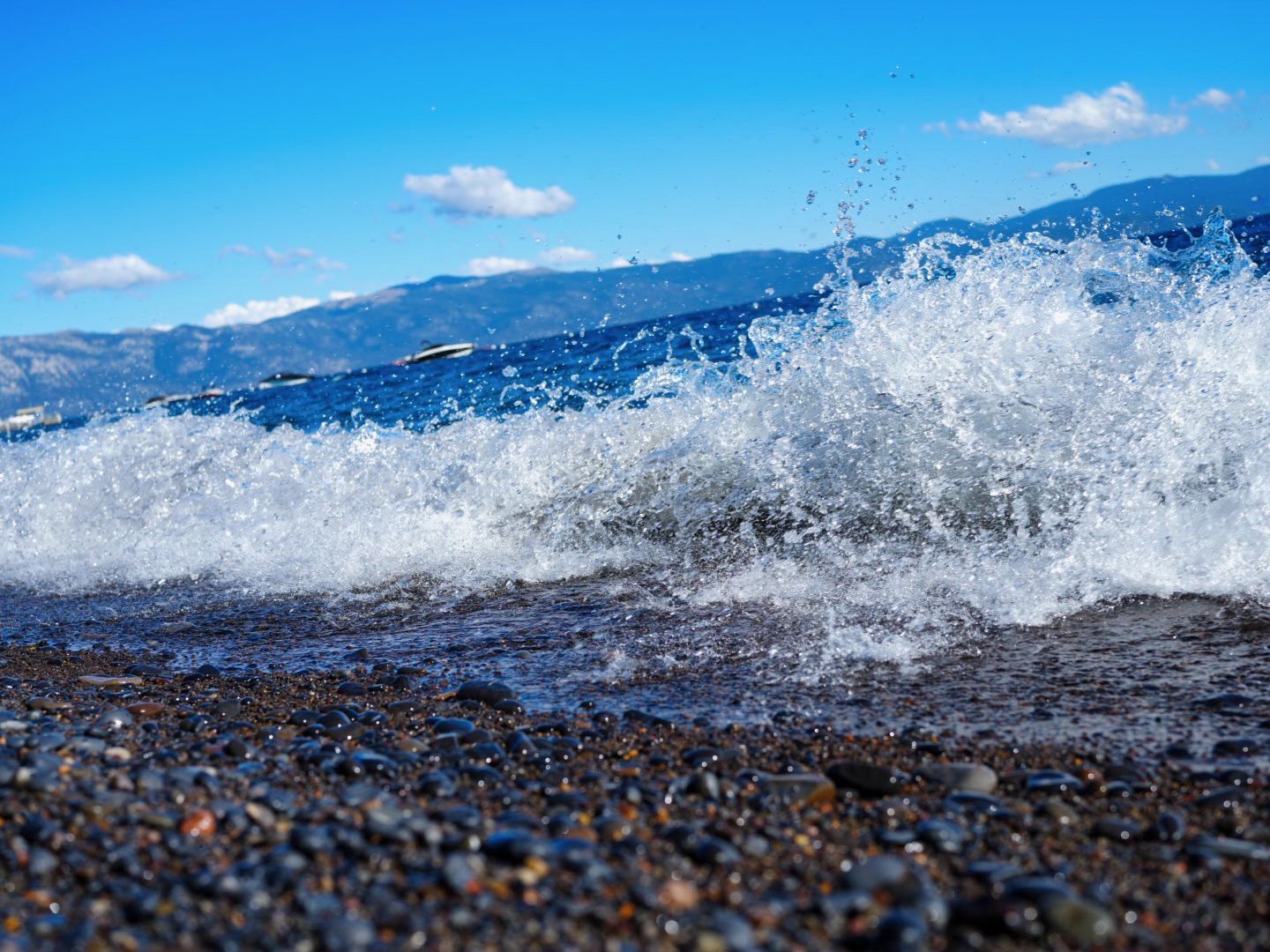 A wave breaking over pebbles, with blue water and mountains beyond